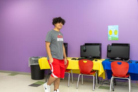 An Eastlake High School student is walking by a row of computers with a purple wall behind him. He is looking towards the camera and smiling.