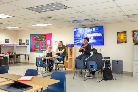  Students and a teacher at Eastlake High School sit together in a classroom. They are looking toward a speaker. There is a large screen behind them. 