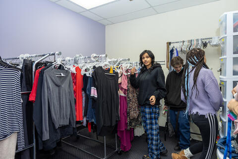 Three students at Eastlake High School browse the community closet, a place to obtain clothing, free of charge. There are racks of clothes neatly organized on hangers and drawers full of items.