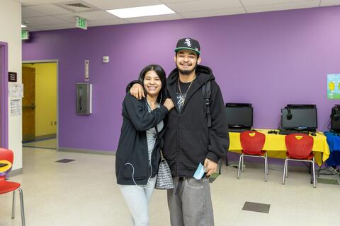 Two students at Eastlake High School pose together for a photo. They are standing in the school in front of a row of computers on tables and a purple wall.