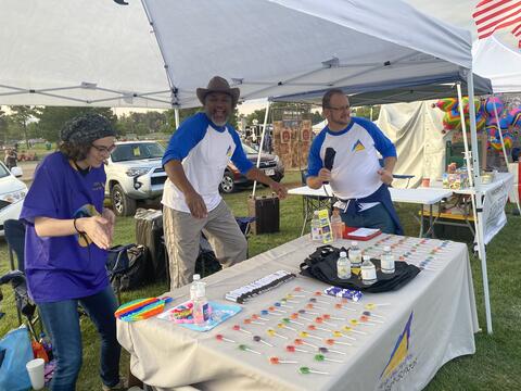 3 members of the Eastlake High School community work and play at a festival booth under a tent. There is a table in front of them with items for the festival attendees. 