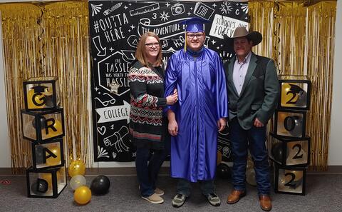 An Eastlake High School graduate and parents stand in front of a Grad 2022 backdrop. The graduate is wearing a traditional, purple and gold cap and gown.