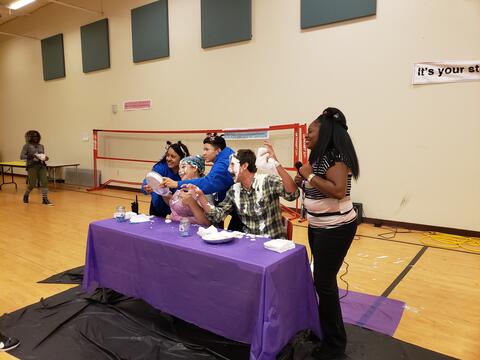 Teachers and students at Eastlake High School pose for a photo after receiving a whipped cream pie in the face.  They are seated at a table with a purple tablecloth in the school gymnasium.
