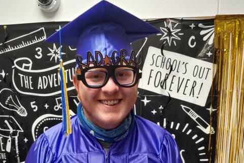 An Eastlake High School 2022 graduate smiles while wearing a purple cap and gown and glasses that say “Rad Grad.”
