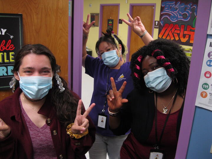  1 Student and 2 Eastlake High School staff members stand together in a classroom doorway. They are smiling under masks and holding up peace fingers.
